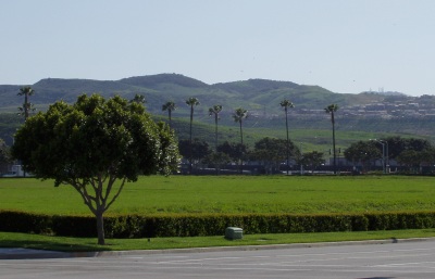 Street corner, facing a vacant lot covered with green grass, in front of green hills with a clutch of houses to the right.